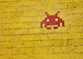 Street art featuring a retro pixelated space invader on a vivid yellow brick wall.